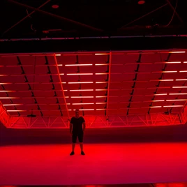 Personne debout sous une grande structure de lumière suspendue, éclairée en rouge dans un espace intérieur sombre