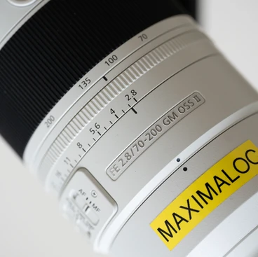 Gros plan sur un objectif Sony 70-200 F2.8 GM II avec les détails des échelles de focale et d'ouverture et une étiquette jaune portant l'inscription 'MAXIMALOC'