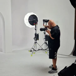 Photographe en tenue noire utilisant un appareil photo professionnel sur trépied, avec un éclairage de studio à parapluie, dans un décor blanc épuré avec des arches